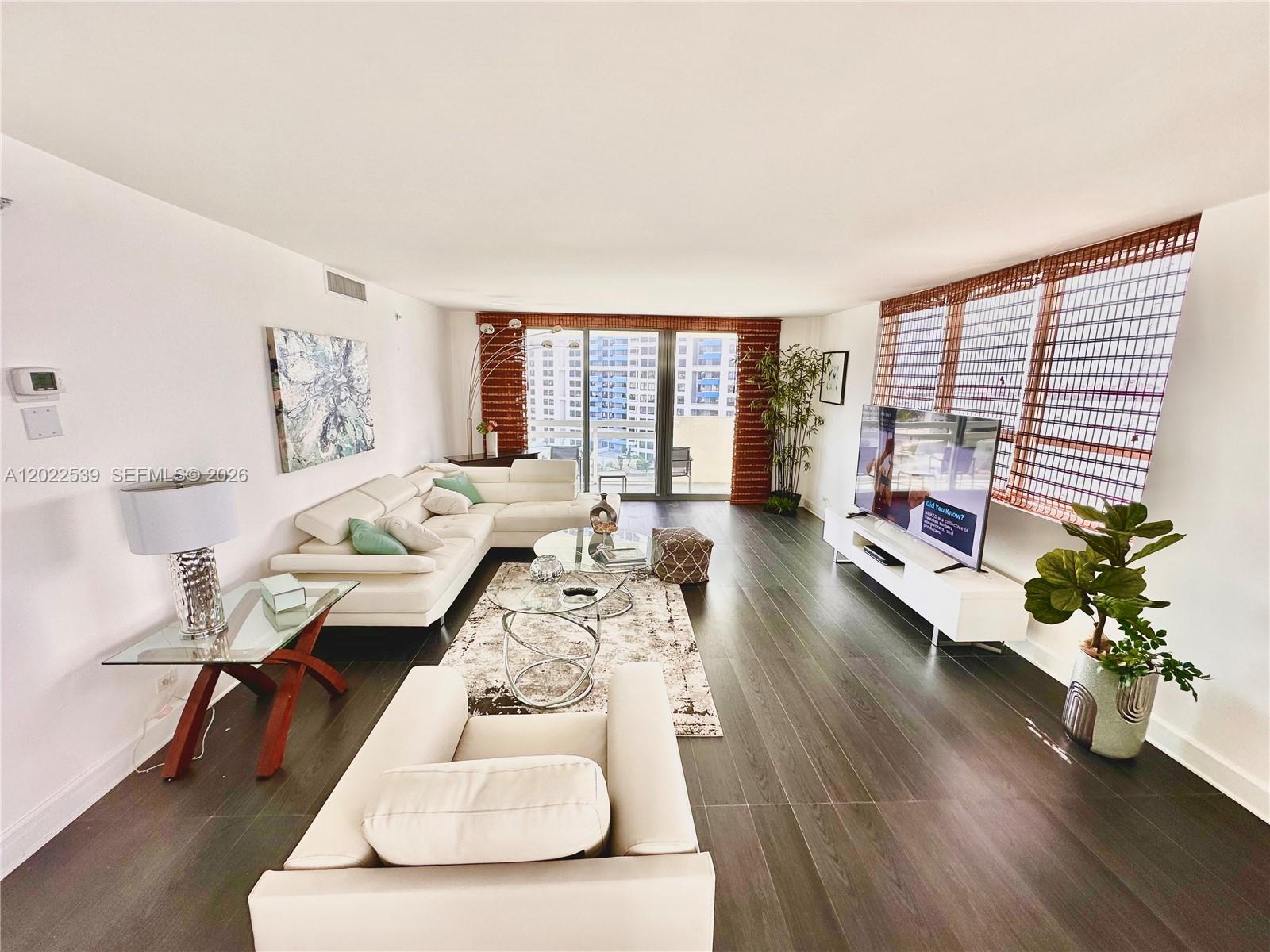 A modern living room features white sofas, dark wood flooring, and large windows with blinds — 1500 Bay Rd # 1114S, Miami Beach FL 33139, MIAMI BEACH, FL