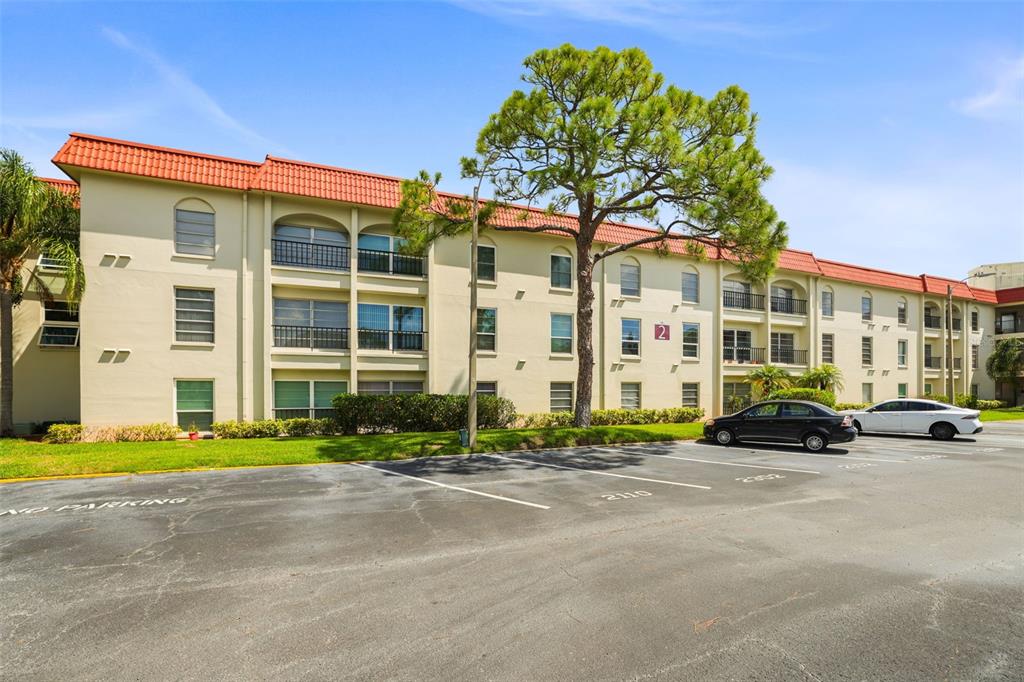 A three-story apartment building with a red tile roof stands behind a paved parking lot — 2700 BAYSHORE BOULEVARD UNIT 2110, DUNEDIN, FL