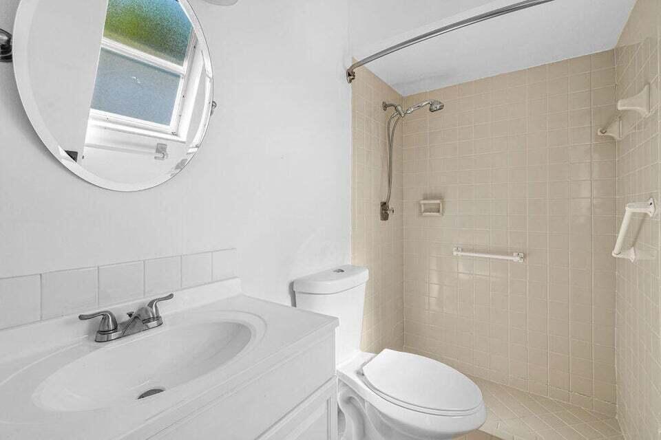 A bathroom features a white vanity, a round mirror, a toilet, and a tiled shower — 9046 E Highland Pines Boulevard E, Palm Beach Gardens FL 33418, PALM BEACH GARDENS, FL