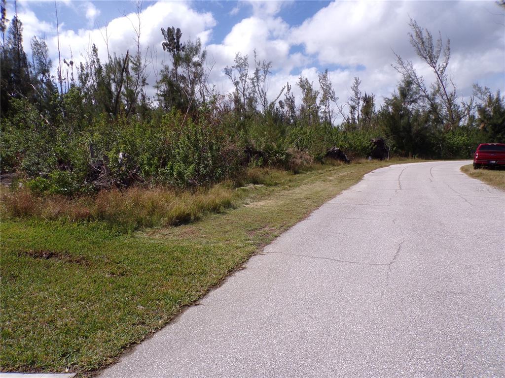 A paved road curves past a vacant, heavily wooded lot under a cloudy sky — 16724 LIGGETT CIRCLE, PORT CHARLOTTE, FL