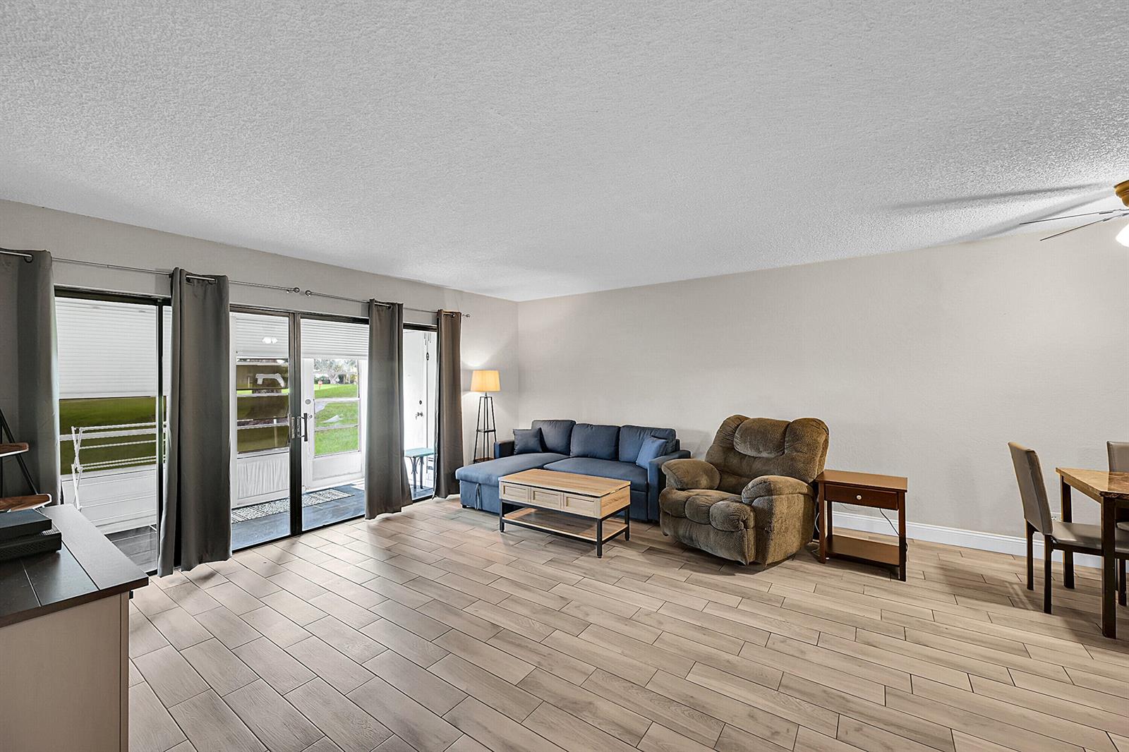 A living room with wood-look tile floors, a blue sectional, and sliding glass patio doors — 497 Piedmont K, Delray Beach FL 33484, DELRAY BEACH, FL