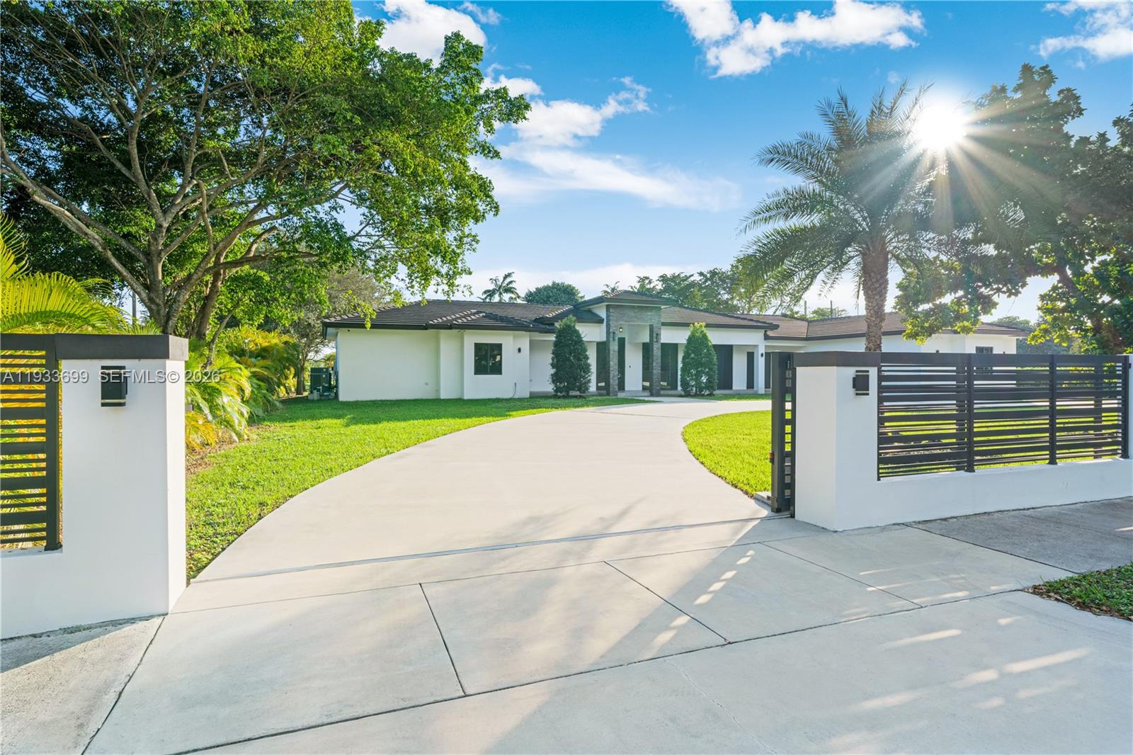 A modern white house with a curved driveway is viewed through an open gated entrance — 10398 SW 64th St, Miami FL 33173, MIAMI, FL