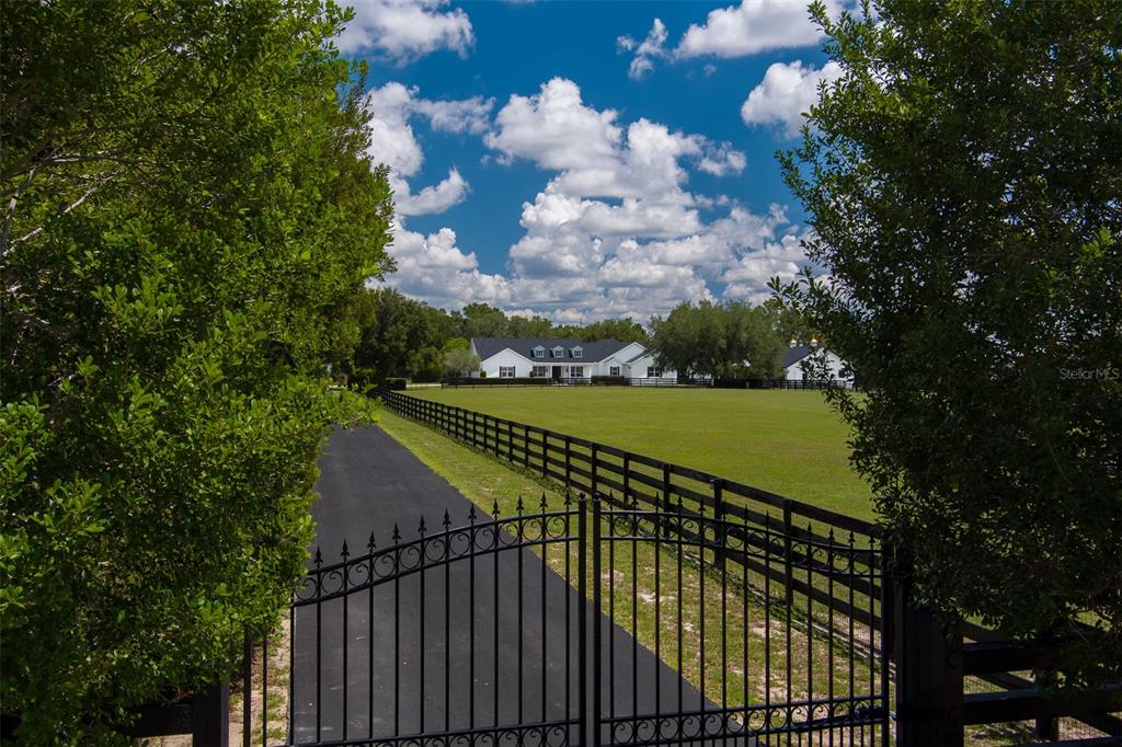 A black metal gate opens to a paved driveway leading toward a white house — 4985 SW 85TH STREET, OCALA, FL