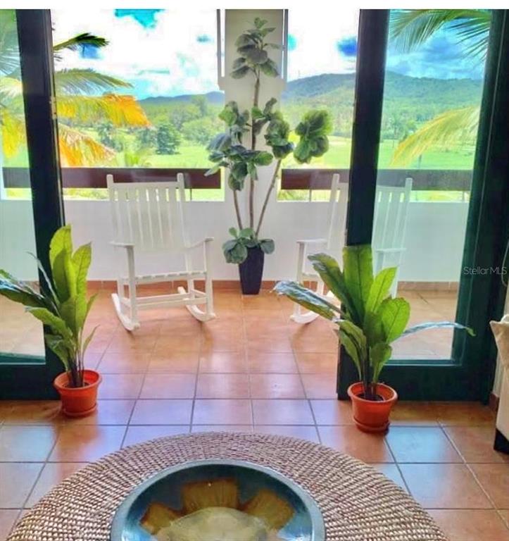 A tiled balcony features two white rocking chairs, potted plants, and a scenic mountain view — 802 RIO MAR VILLAGE, RIO GRANDE, PR