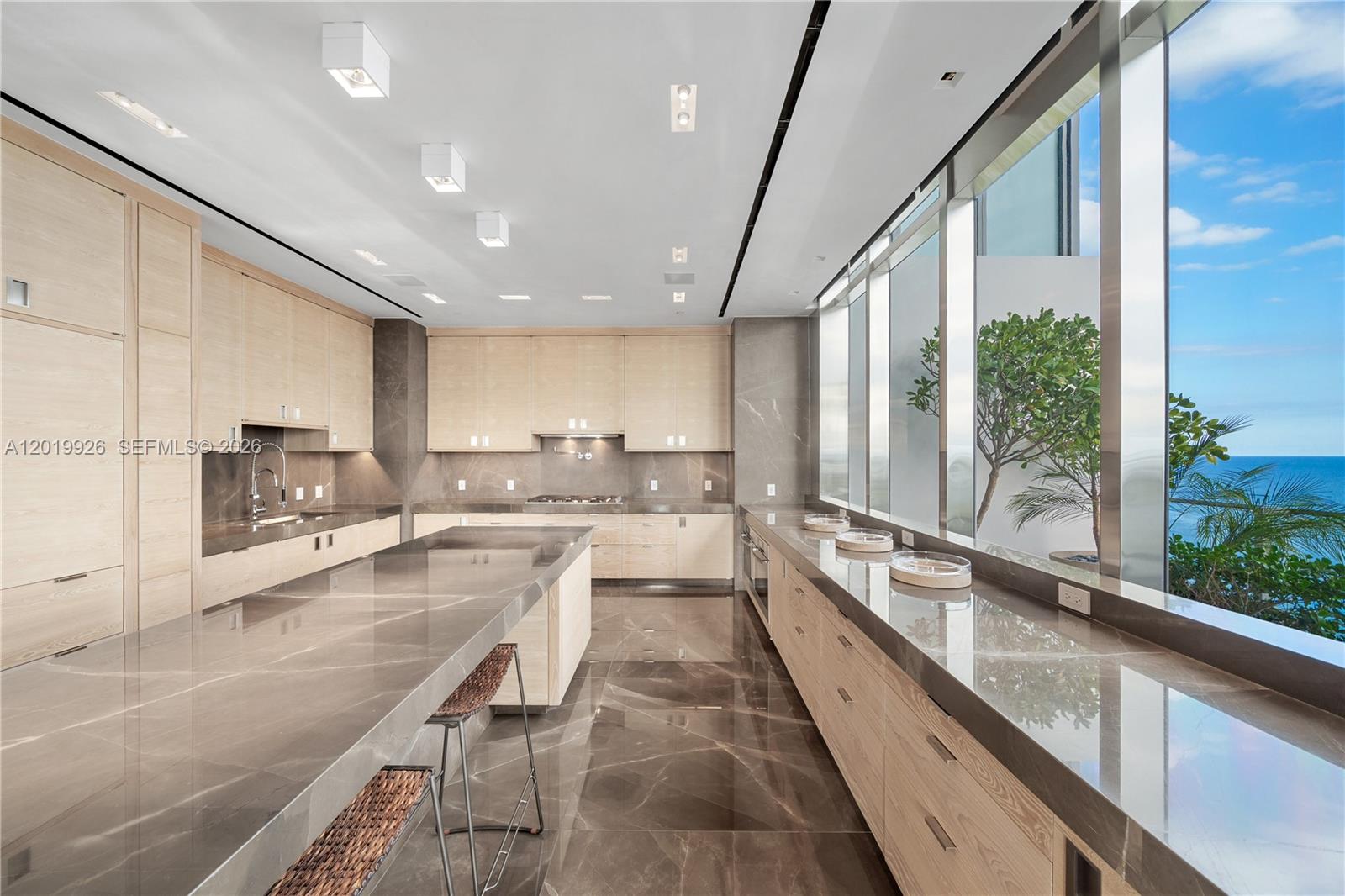 A modern kitchen features light wood cabinetry, dark stone countertops, and floor-to-ceiling windows overlooking the ocean — 800 S Pointe Dr # 2204, Miami Beach FL 33139, MIAMI BEACH, FL