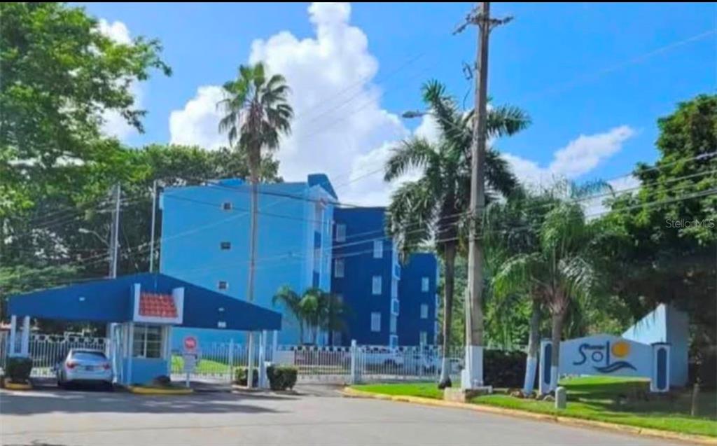 A gated entrance with a blue guardhouse leads to a multi-story blue apartment building — Apartments MONTESOL UNIT E-312, FAJARDO, PR
