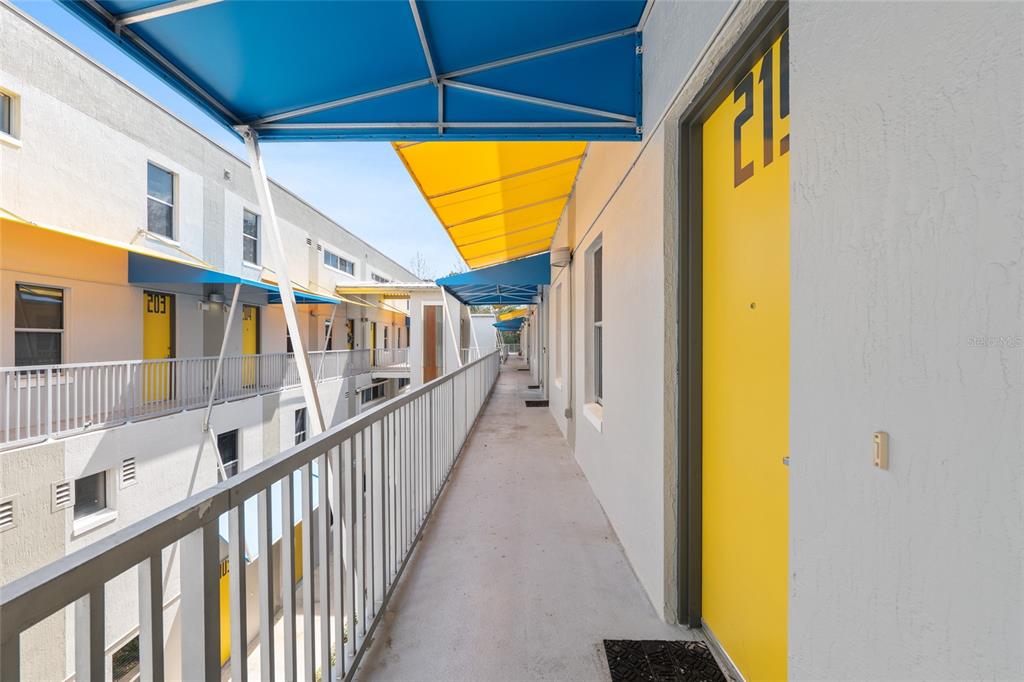 An outdoor walkway with yellow doors and blue awnings overlooks a multi-story apartment complex courtyard — 2515 SW 35TH PLACE UNIT 219, GAINESVILLE, FL