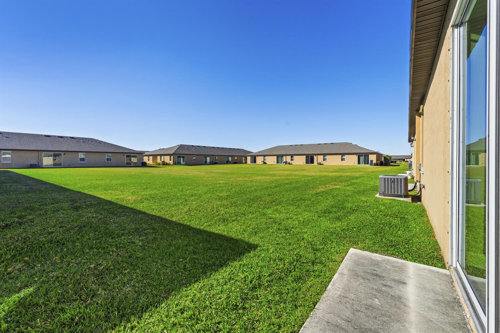 A view of a large, grassy backyard behind a tan house with a concrete patio — 1817 Party Place, Fort Pierce FL 34947, FORT PIERCE, FL