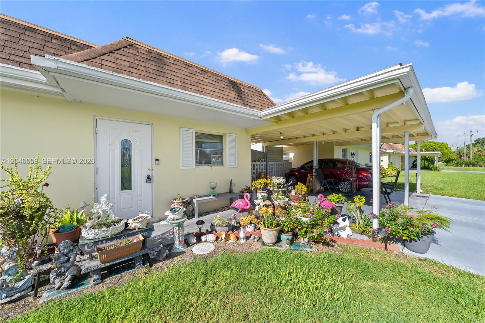 A yellow house exterior features a front door, a carport, and a garden with decorations — 2208 Gardenia Way, Lehigh Acres FL 33936, LEHIGH ACRES, FL