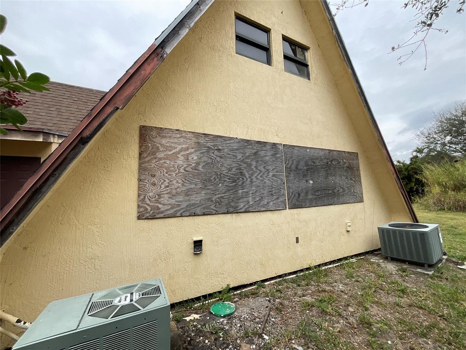 An exterior view of a yellow A-frame house with boarded-up windows and two AC units — 6650 Riggers Road, Lake Worth FL 33462, LAKE WORTH, FL