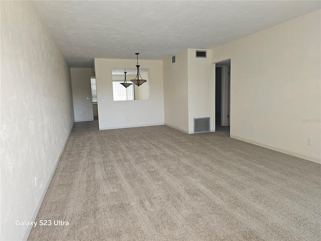 An empty living room features beige carpet, off-white walls, and a dining area with a chandelier — 2454 AUSTRALIA WAY E UNIT 46, CLEARWATER, FL