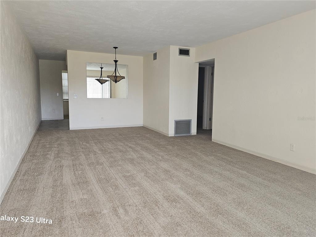 An empty living room features beige carpeting, off-white walls, a dining area mirror, and a chandelier — 2454 AUSTRALIA WAY E UNIT 46, CLEARWATER, FL