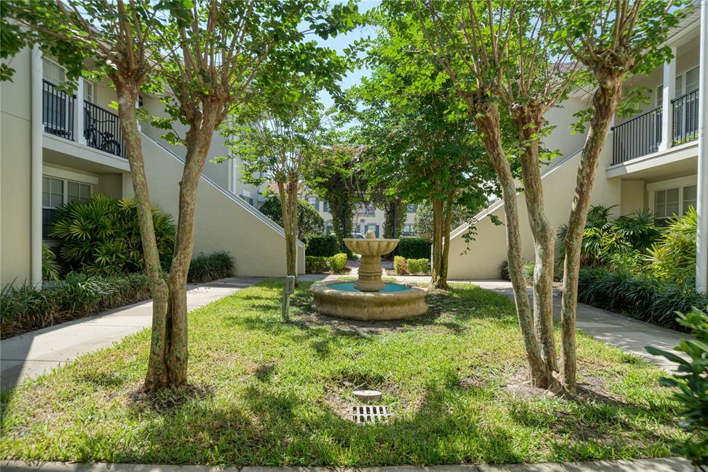 A courtyard between two apartment buildings features a stone fountain surrounded by grass and trees — 714 SIENA PALM DRIVE UNIT 104, CELEBRATION, FL