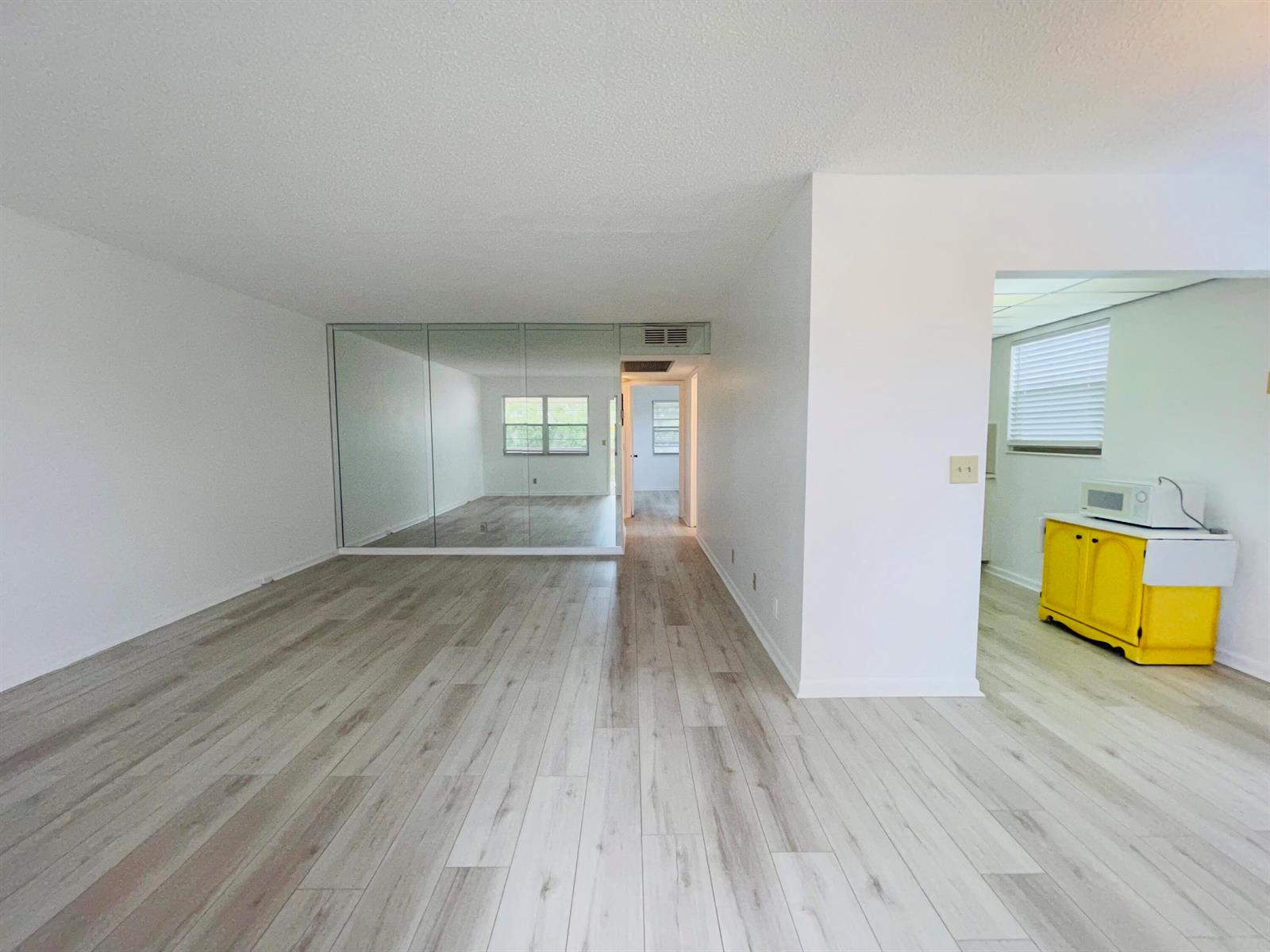 A living room with light wood-look flooring, a mirrored wall, and an adjacent kitchen area — 333 Farnham P, Deerfield Beach FL 33442, DEERFIELD BEACH, FL