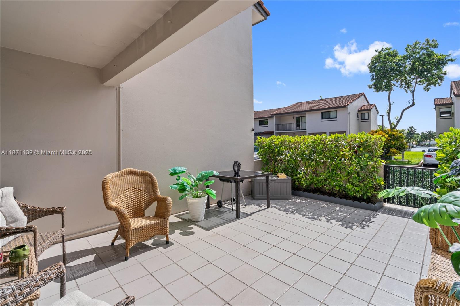 A tiled patio features wicker furniture, a small table, and a hedge against a blue sky — 13412 SW 62nd St # 103M, Miami FL 33183, MIAMI, FL