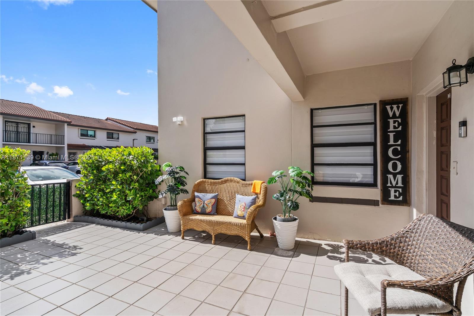 A paved patio features a wicker bench, potted plants, and a welcome sign by the entrance — 13412 SW 62nd St # 103M, Miami FL 33183, MIAMI, FL