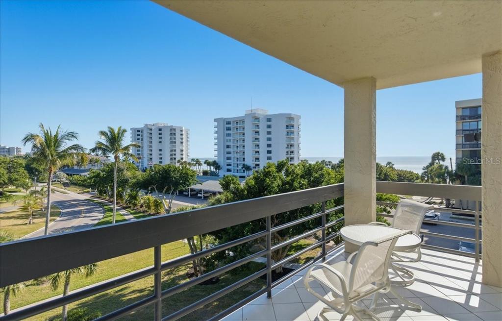 A balcony with white patio furniture overlooks a coastal landscape with palm trees and buildings — 1065 GULF OF MEXICO DRIVE UNIT 305, LONGBOAT KEY, FL