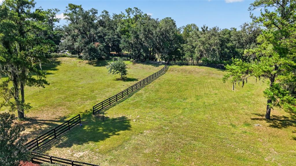 An aerial view shows a grassy pasture enclosed by black wooden fencing and mature trees — 11050 SE 37TH AVENUE ROAD, BELLEVIEW, FL