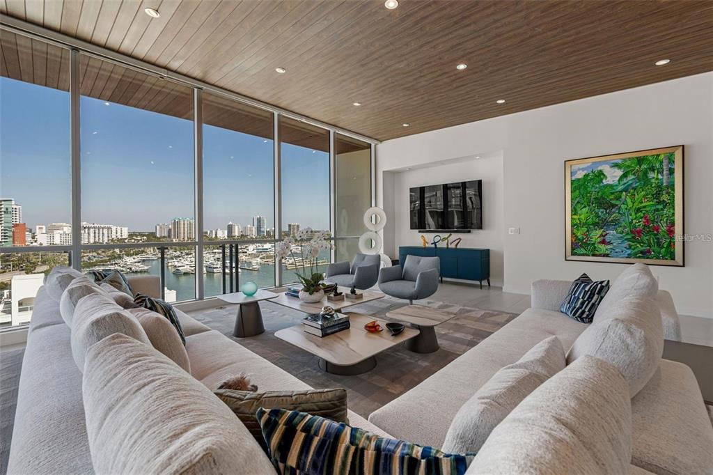 A modern living room features a wood-paneled ceiling, floor-to-ceiling windows, and a harbor view — 111 GOLDEN GATE POINT UNIT 802PH, SARASOTA, FL