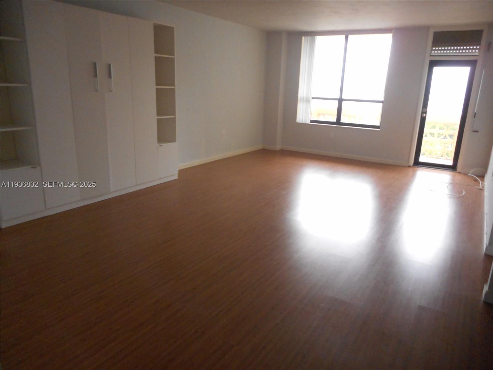 An empty room features wood-look flooring, white built-in cabinetry, a large window, and a balcony door — 10185 Collins Ave # 1204, Bal Harbour FL 33154, BAL HARBOUR, FL