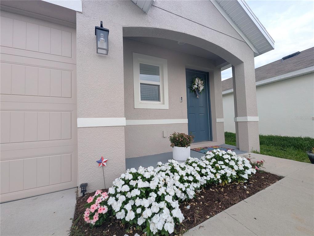 A beige house exterior features a blue front door, a garage, and white flower beds — 37377 ANGEL WINGS AVENUE, ZEPHYRHILLS, FL