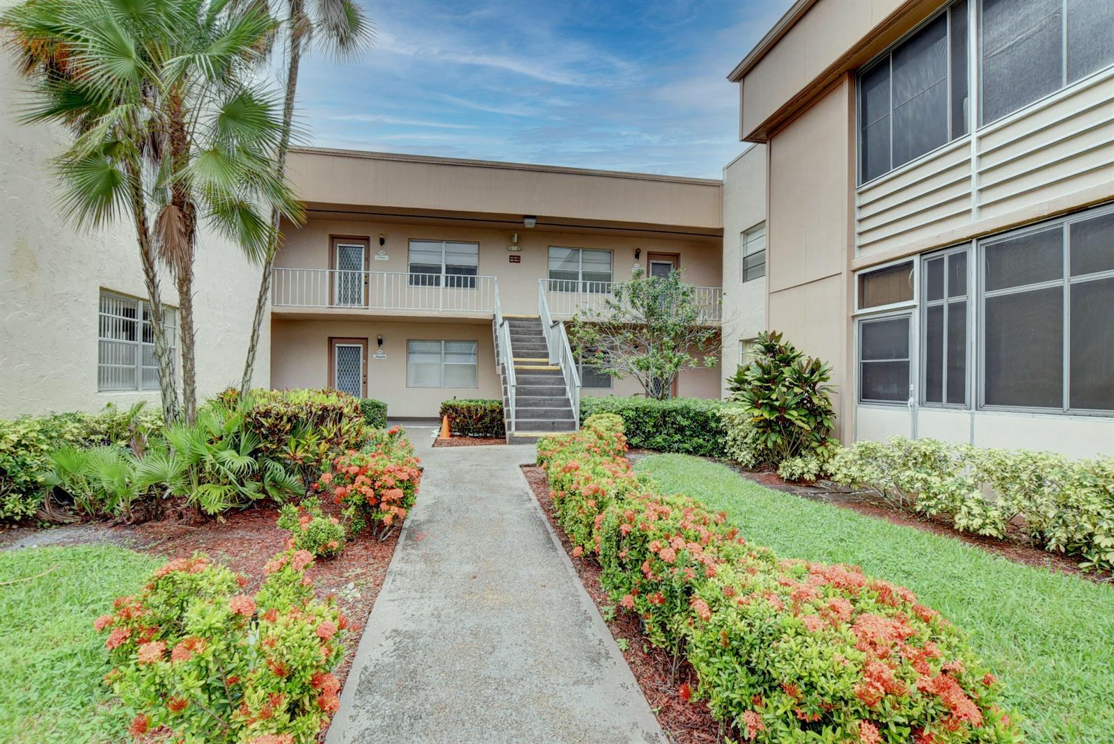 A concrete walkway leads to a two-story apartment building with stairs and landscaped gardens — 138 Flanders C, Delray Beach FL 33484, DELRAY BEACH, FL