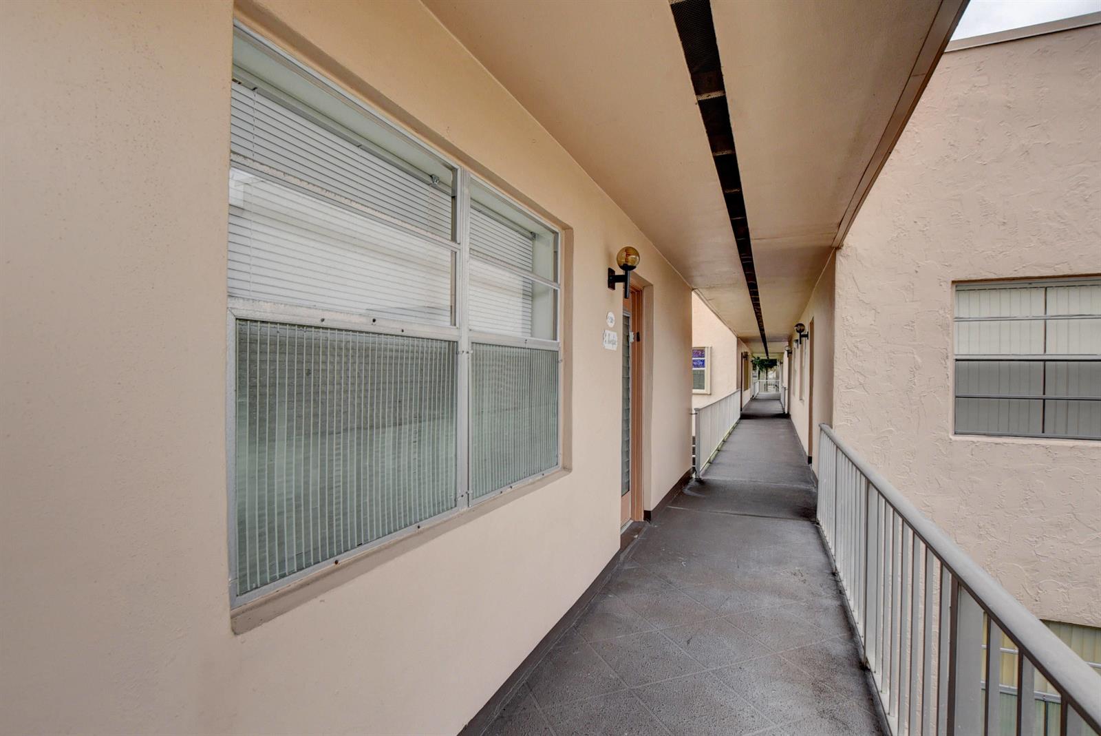 An outdoor apartment building walkway with beige walls, a gray floor, and metal railings — 138 Flanders C, Delray Beach FL 33484, DELRAY BEACH, FL