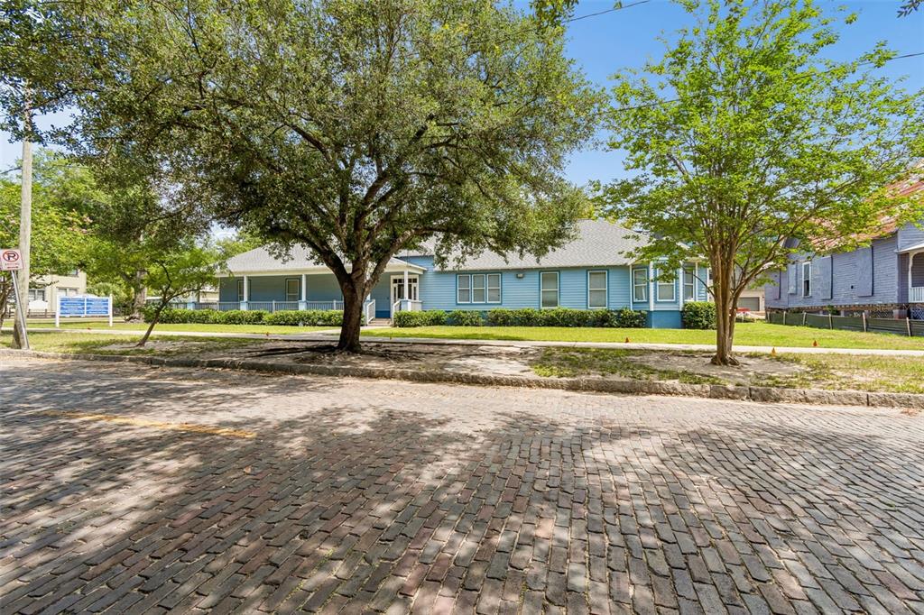 A blue single-story house with a wrap-around porch sits behind a large tree and cobblestone street — 402 E 7TH AVENUE, TAMPA, FL