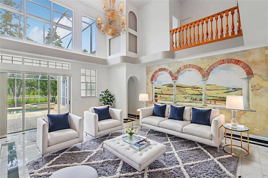 A bright, high-ceilinged living room features a large wall mural, white furniture, and a chandelier — 3062 SUTTON WOODS DRIVE, PLANT CITY, FL