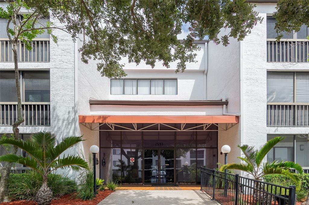 The exterior of a multi-story apartment building features a glass entrance under an orange awning — 2593 COUNTRYSIDE BOULEVARD UNIT 7210, CLEARWATER, FL