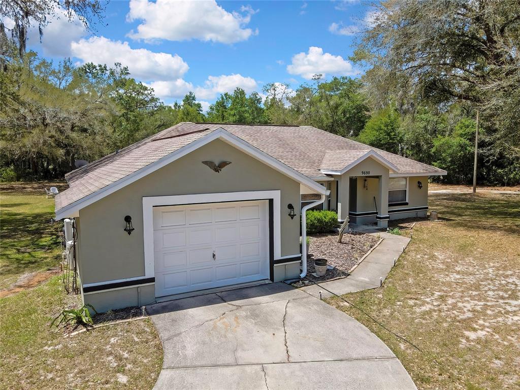 An exterior view of a single-story house with a garage, concrete driveway, and wooded surroundings — 9690 N ALDER AVENUE, CRYSTAL RIVER, FL