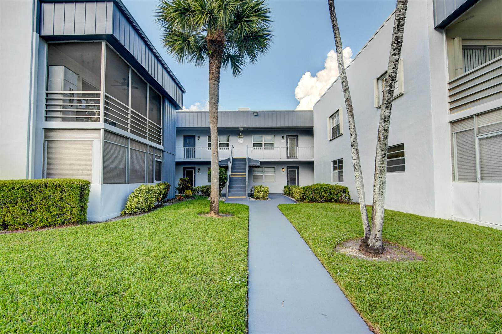 A paved walkway leads to the entrance of a two-story apartment building with palm trees — 42 Burgundy A # A, Delray Beach FL 33484, DELRAY BEACH, FL