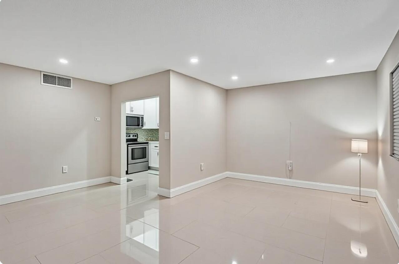 An empty room with light-colored tile flooring, beige walls, and an opening to a kitchen — 4980 E Sabal Palm Boulevard E # 124, Tamarac FL 33319, TAMARAC, FL