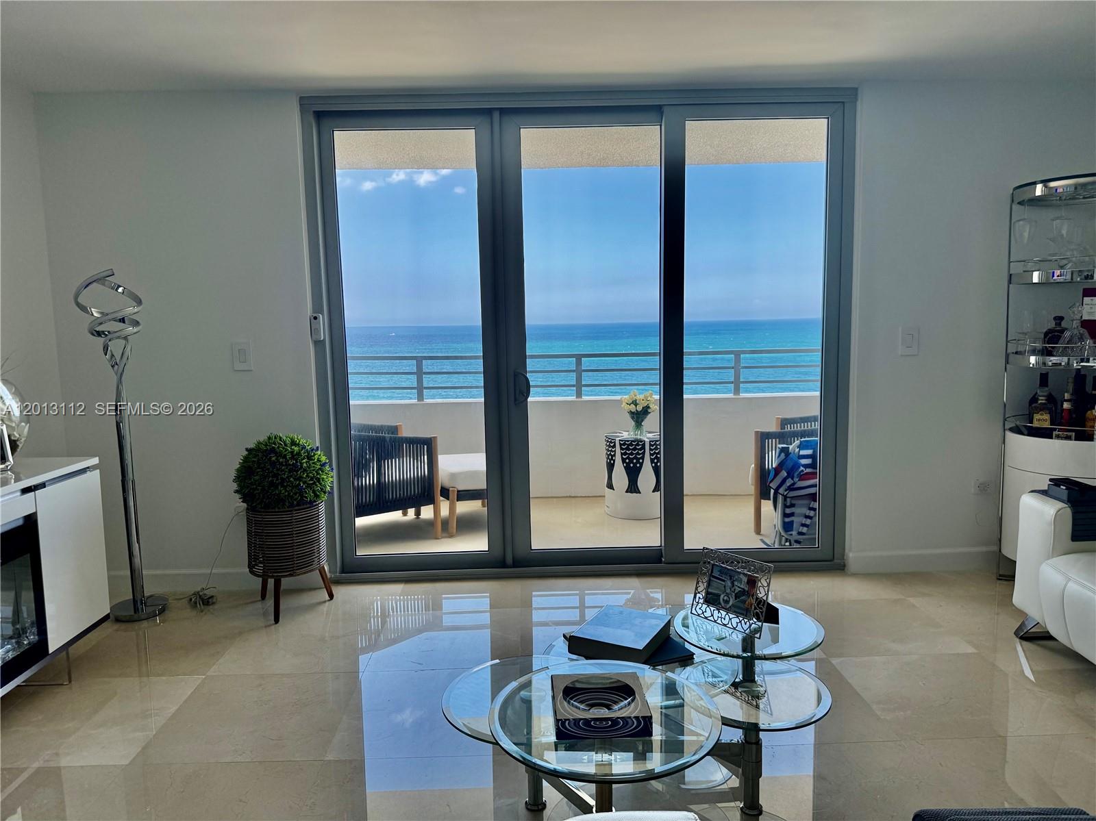 A living room features a glass coffee table and sliding doors opening to an oceanfront balcony — 5555 Collins Ave # 14E, Miami Beach FL 33140, MIAMI BEACH, FL