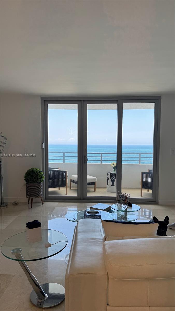 A living room with a white sofa, glass tables, and sliding doors opening to an oceanfront balcony — 5555 Collins Ave # 14E, Miami Beach FL 33140, MIAMI BEACH, FL