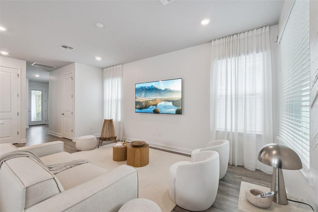 A bright living room features a wall-mounted television, white furniture, and light wood flooring — 6935 HIGGS ALLEY, ORLANDO, FL