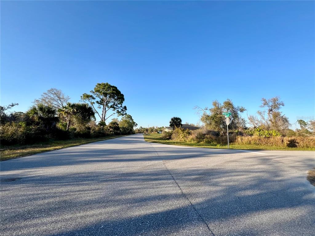 A paved road stretches into the distance, flanked by trees and brush under a clear sky — 14113 MAGNESS AVENUE, PORT CHARLOTTE, FL