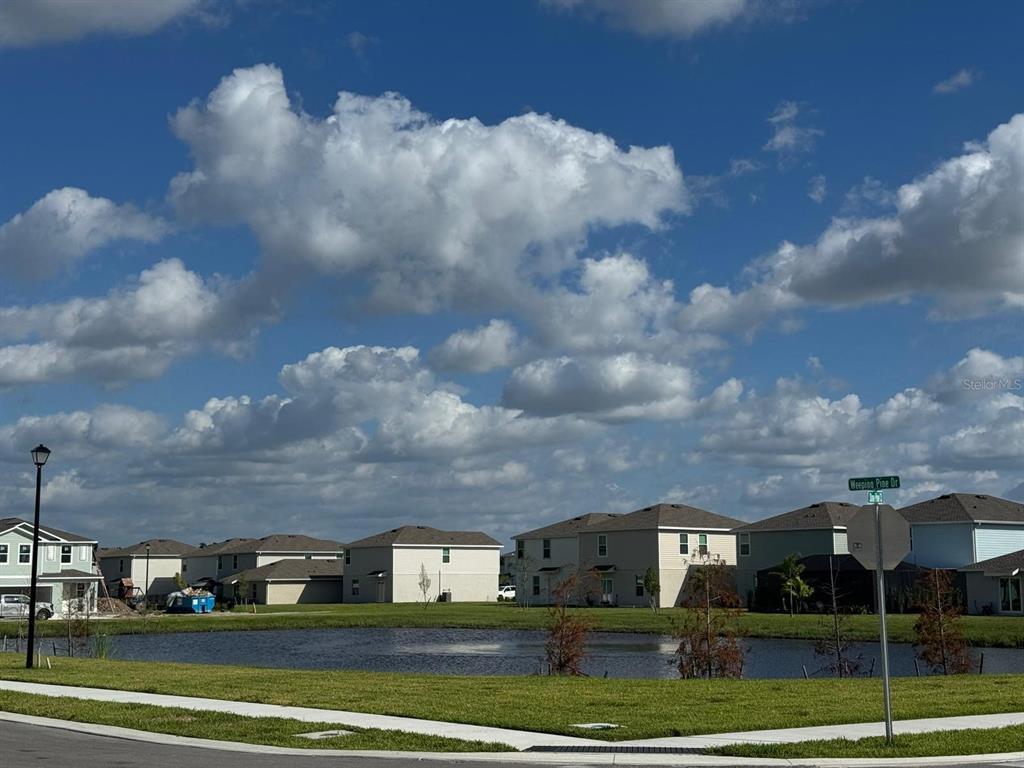 A pond sits in front of several suburban houses under a blue sky with clouds — 7111 WEEPING PINE DRIVE, FORT MYERS, FL