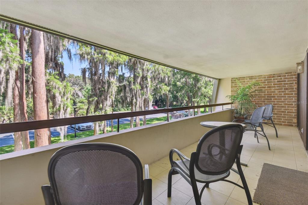 A covered balcony with a brick wall, tiled floor, and outdoor chairs overlooking wooded grounds — 36750 US HIGHWAY 19 N UNIT 04239, PALM HARBOR, FL