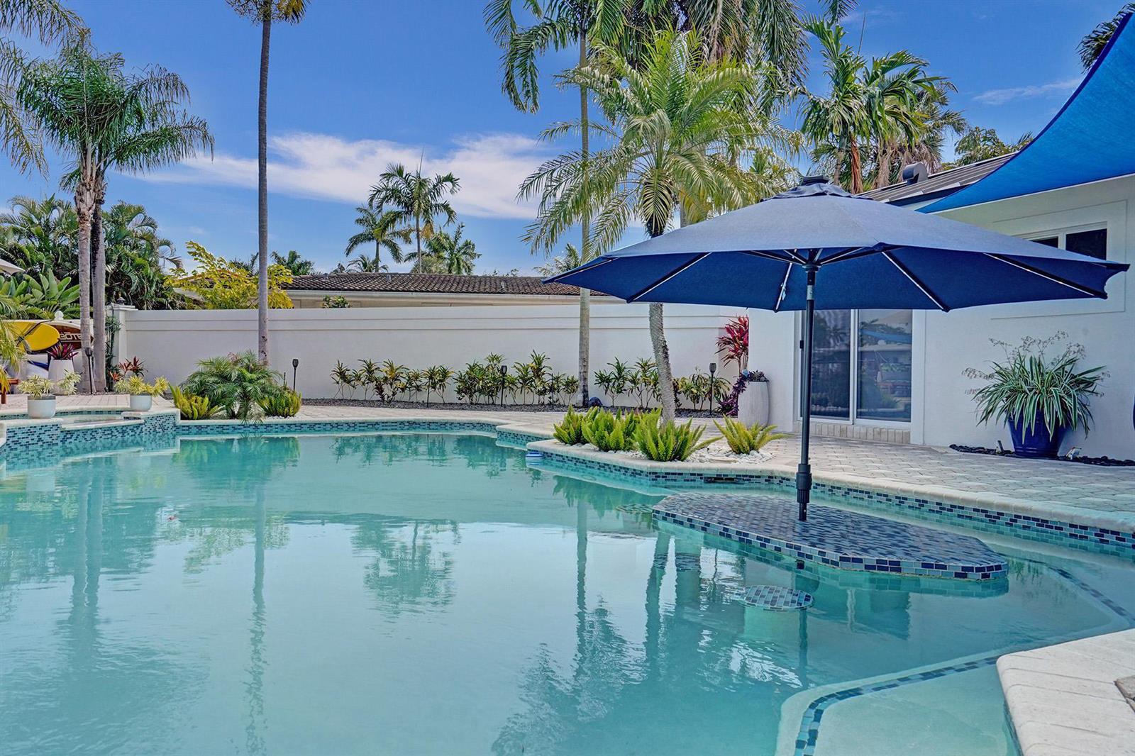 A swimming pool with a built-in table and blue umbrella sits beside a white house — 201 SE 11th Street, Pompano Beach FL 33060, POMPANO BEACH, FL
