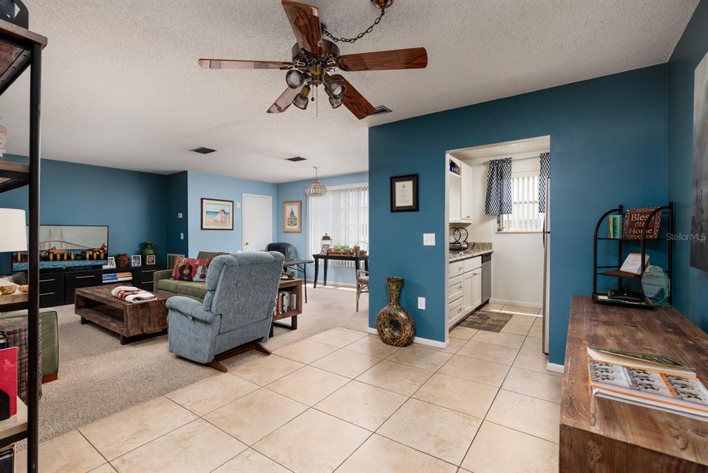 A living room with blue walls, a ceiling fan, and tile flooring leads to a kitchen — 5623 80TH STREET N UNIT 504, ST PETERSBURG, FL
