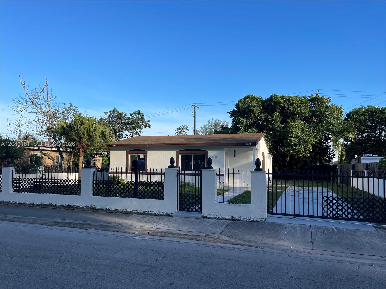 A single-story white house with a brown roof is enclosed by a black metal fence — 1310 Sharazad Blvd, Opa-Locka FL 33054, OPA-LOCKA, FL