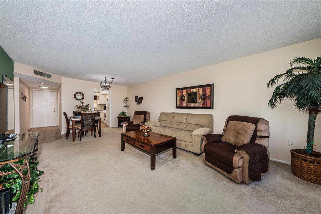 A living room with beige carpet, a tan sofa, two brown armchairs, and a dining area — 4725 COVE CIRCLE UNIT 210, ST PETERSBURG, FL