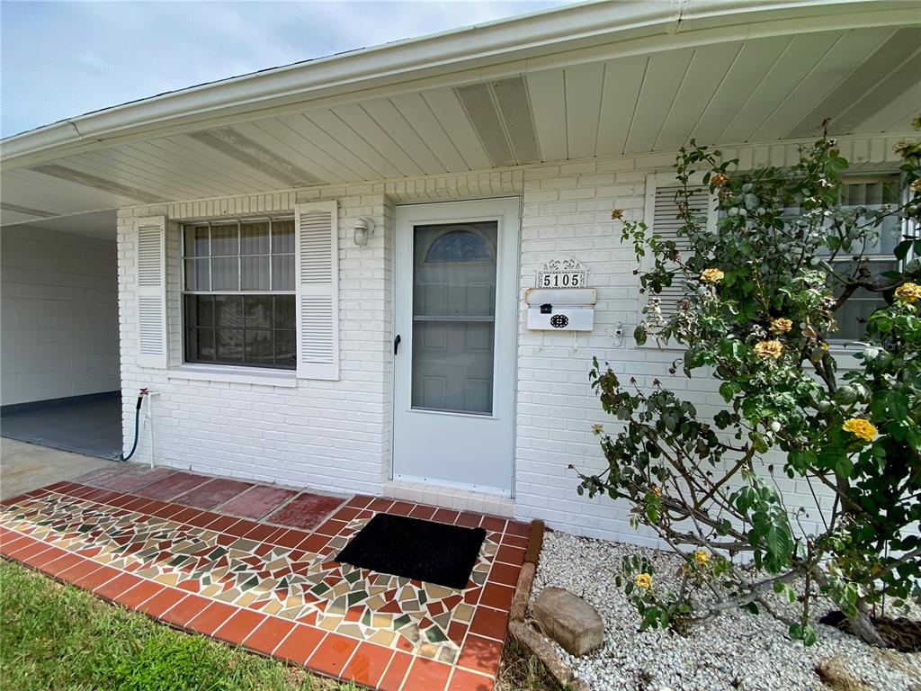 A white brick house exterior features a front door, a tiled porch, and a rose bush — 5105 ROSE PLACE N, PINELLAS PARK, FL