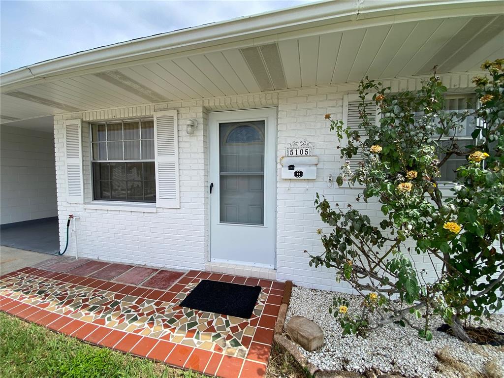 A white brick house exterior features a mosaic-tiled porch, a storm door, and a rose bush — 5105 ROSE PLACE N, PINELLAS PARK, FL