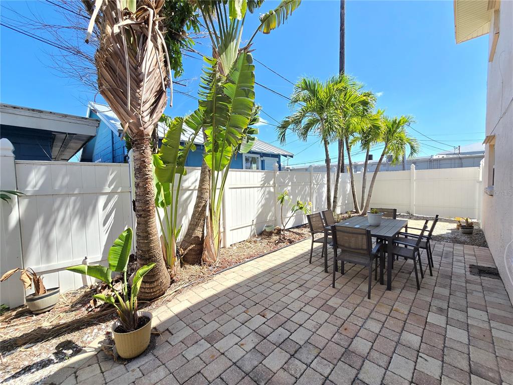 A paved patio features a dining set, potted plants, and palm trees against a white fence — 525 73RD AVENUE UNIT 2, ST PETE BEACH, FL