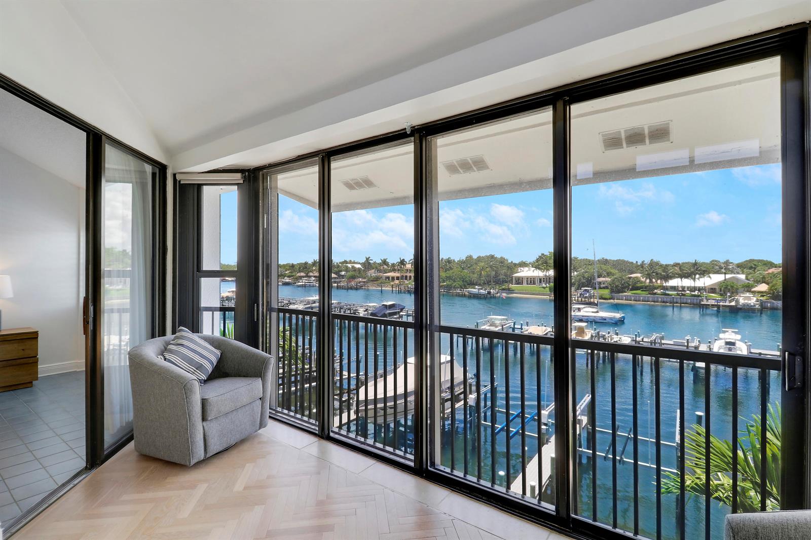 A sunroom with herringbone wood floors and a gray armchair overlooks a canal with boats — 543 Bay Colony Drive N Slip 8, Juno Beach FL 33408, JUNO BEACH, FL