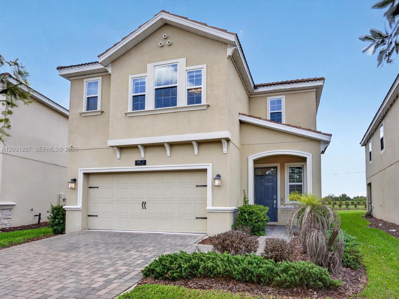 A two-story beige stucco house features a two-car garage, a blue front door, and a paver driveway — 1152 Trappers Trail Loop, Davenport FL 33896, DAVENPORT, FL