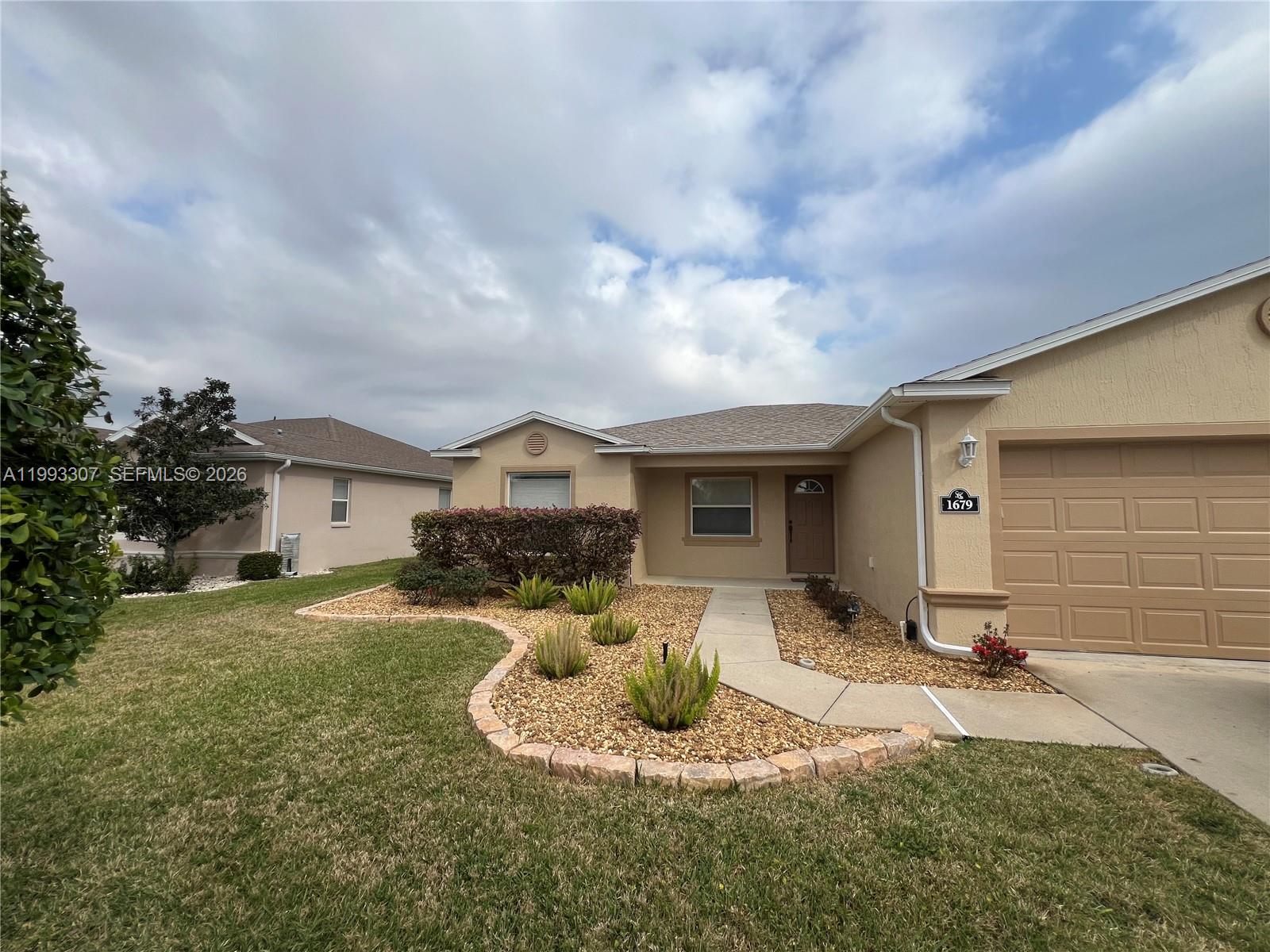 A beige single-story house features a tan garage, a stone-bordered garden, and a concrete walkway — 1679 SW 156th Ln, Ocala FL 34473, OCALA, FL