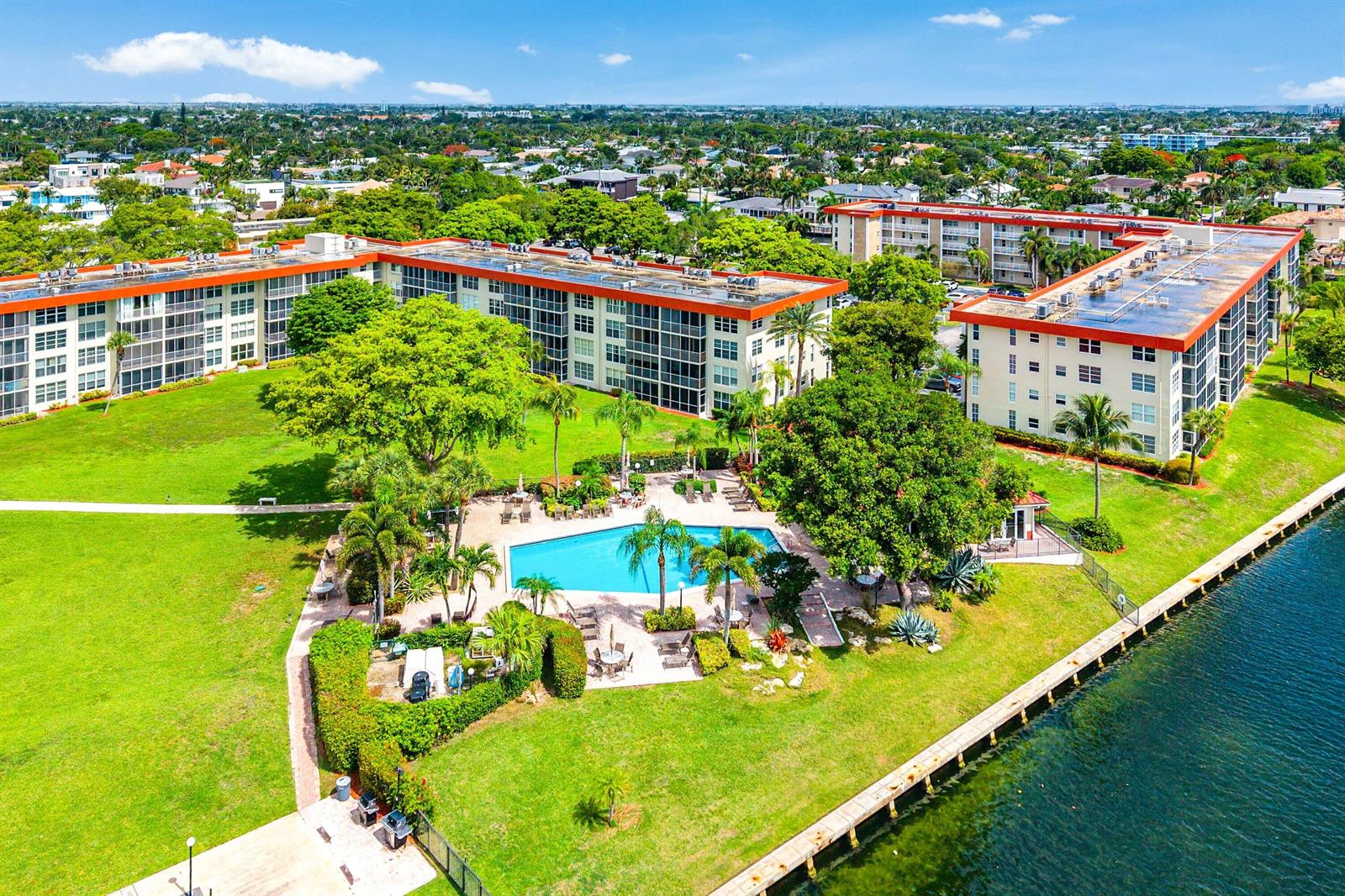 Aerial view of apartment buildings with a swimming pool, green lawn, and waterfront bulkhead — 3150 NE 48th Court # 411, Lighthouse Point FL 33064, LIGHTHOUSE POINT, FL